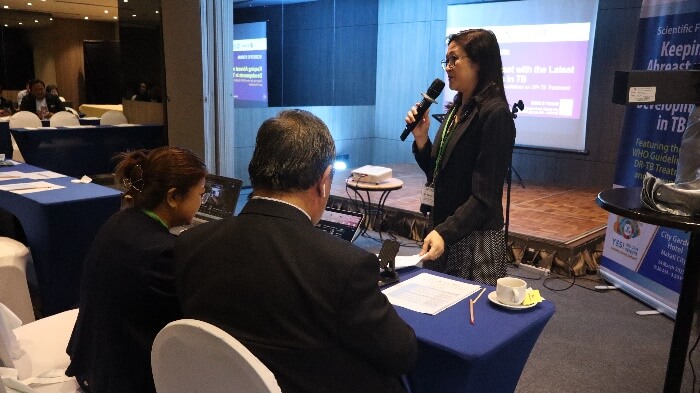 An active seminar moment captures a professional female speaker standing at the front of a meeting room, confidently presenting into a microphone to an attentive, seated listener.