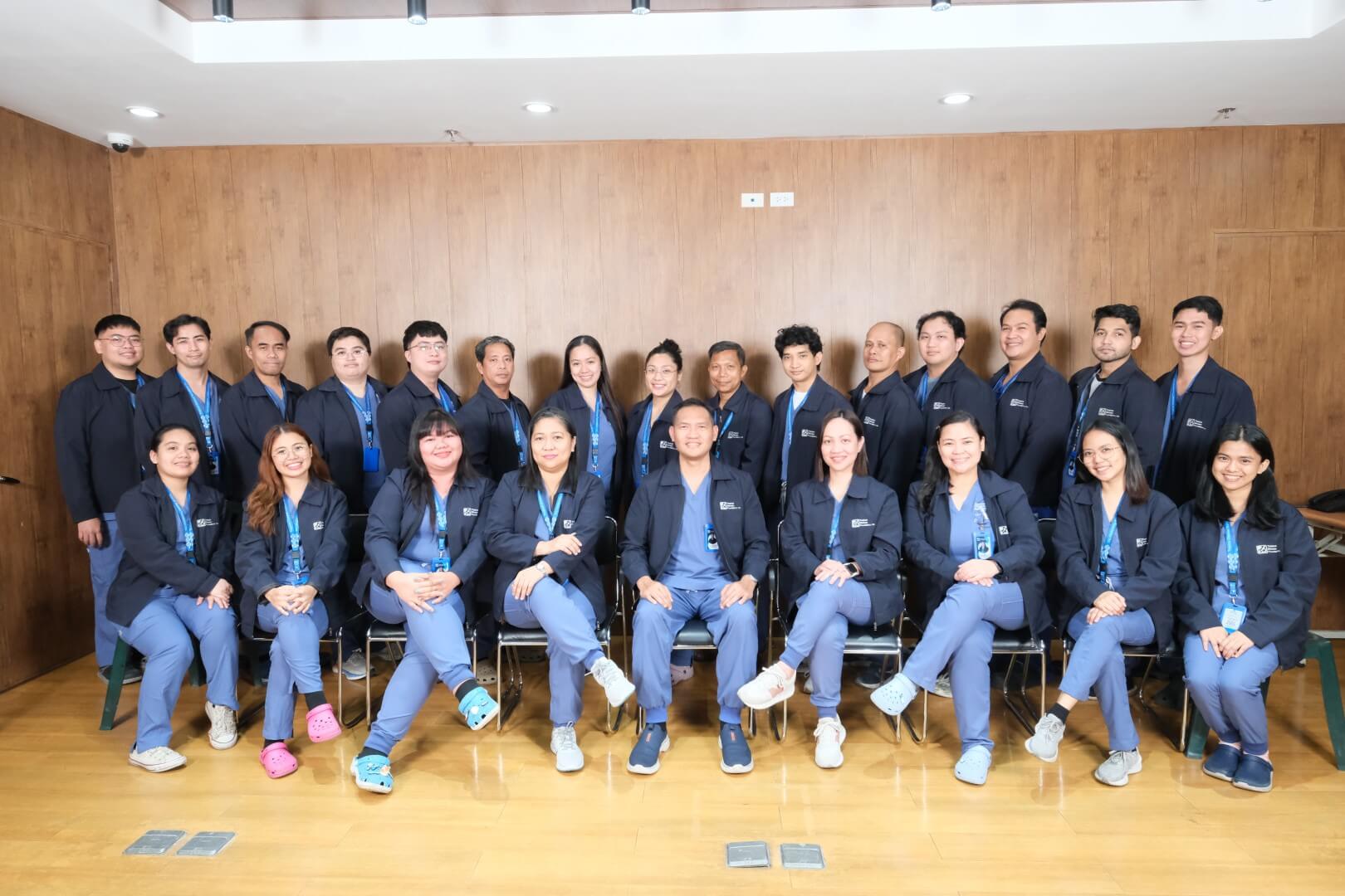 A wide group portrait of medical staff members arranged in two rows. They wear matching dark blue jackets and trousers, smiling together against a polished wooden wall background.