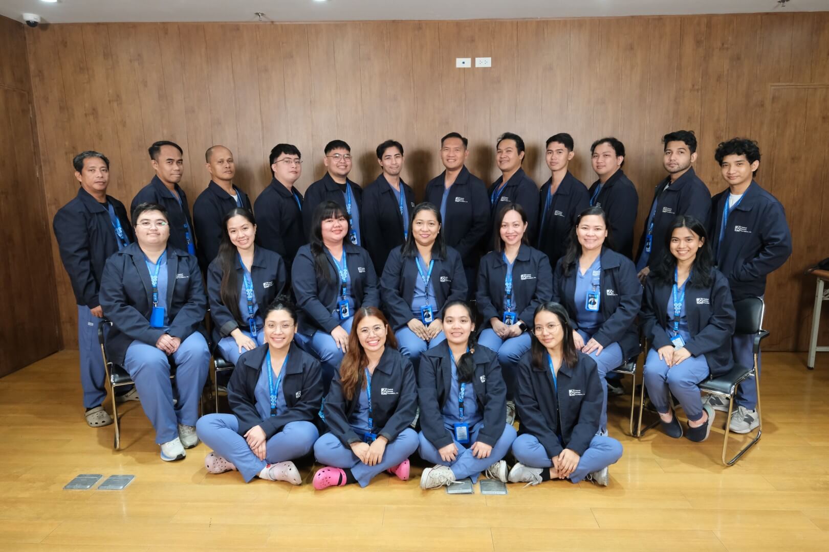 A large team of healthcare workers poses for a group photo in a gym or studio. They are organized in two rows, all wearing matching dark blue uniforms.