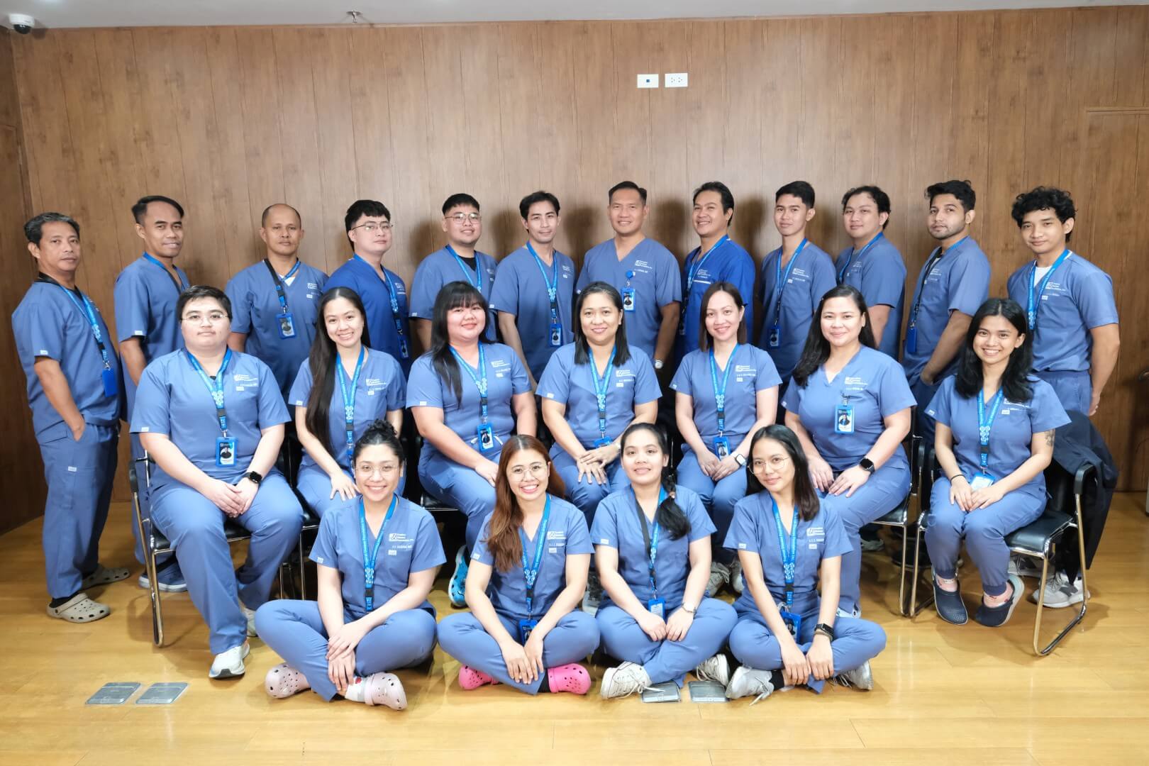 A large group of nurses or clinical staff posing together indoors. Arranged in rows, they wear bright blue medical scrubs and smile warmly for a professional team photo.