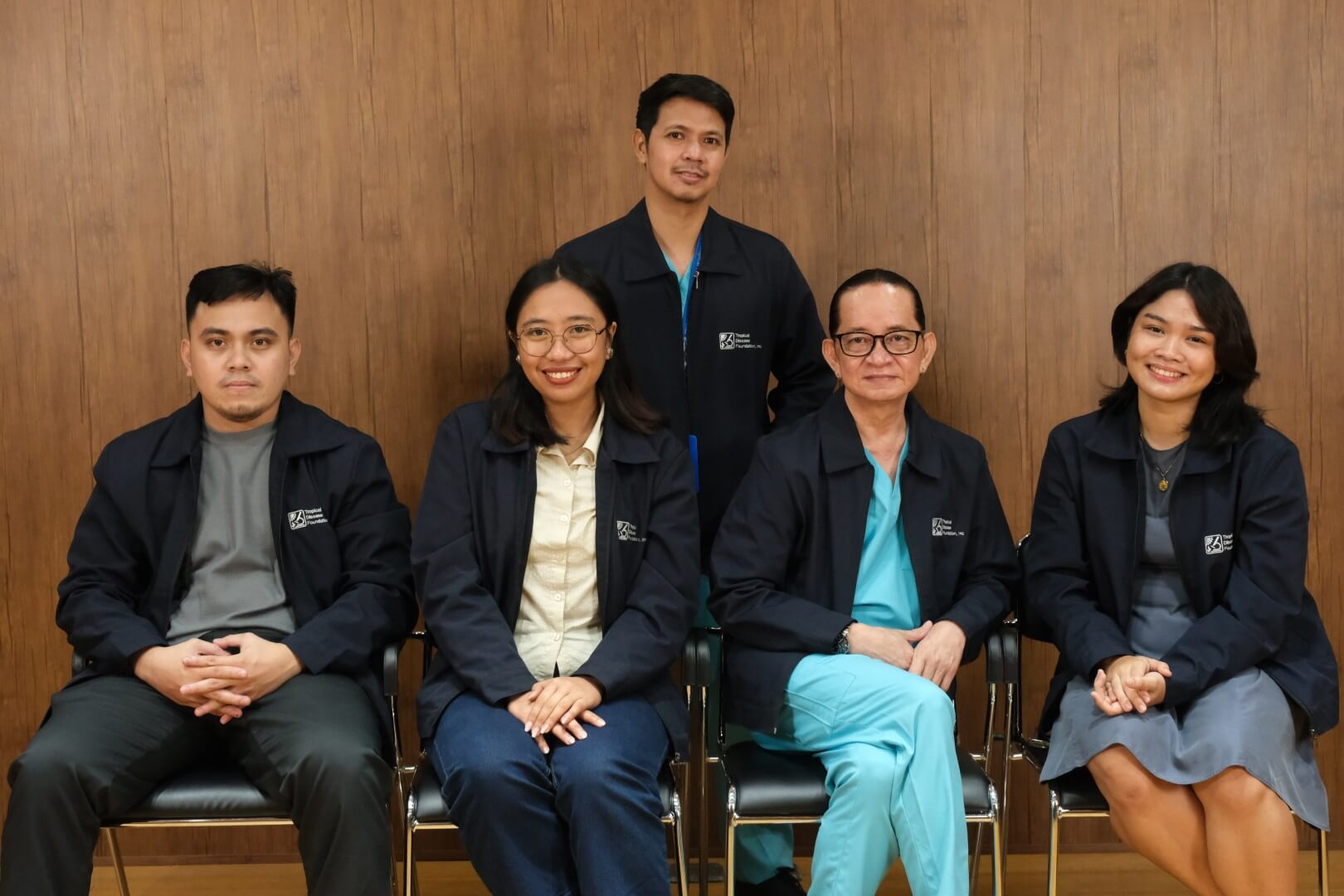 Group portrait of five clinical professionals arranged against a wooden wall backdrop. They wear light teal scrubs and dark corporate jackets, smiling warmly for the official staff photo.