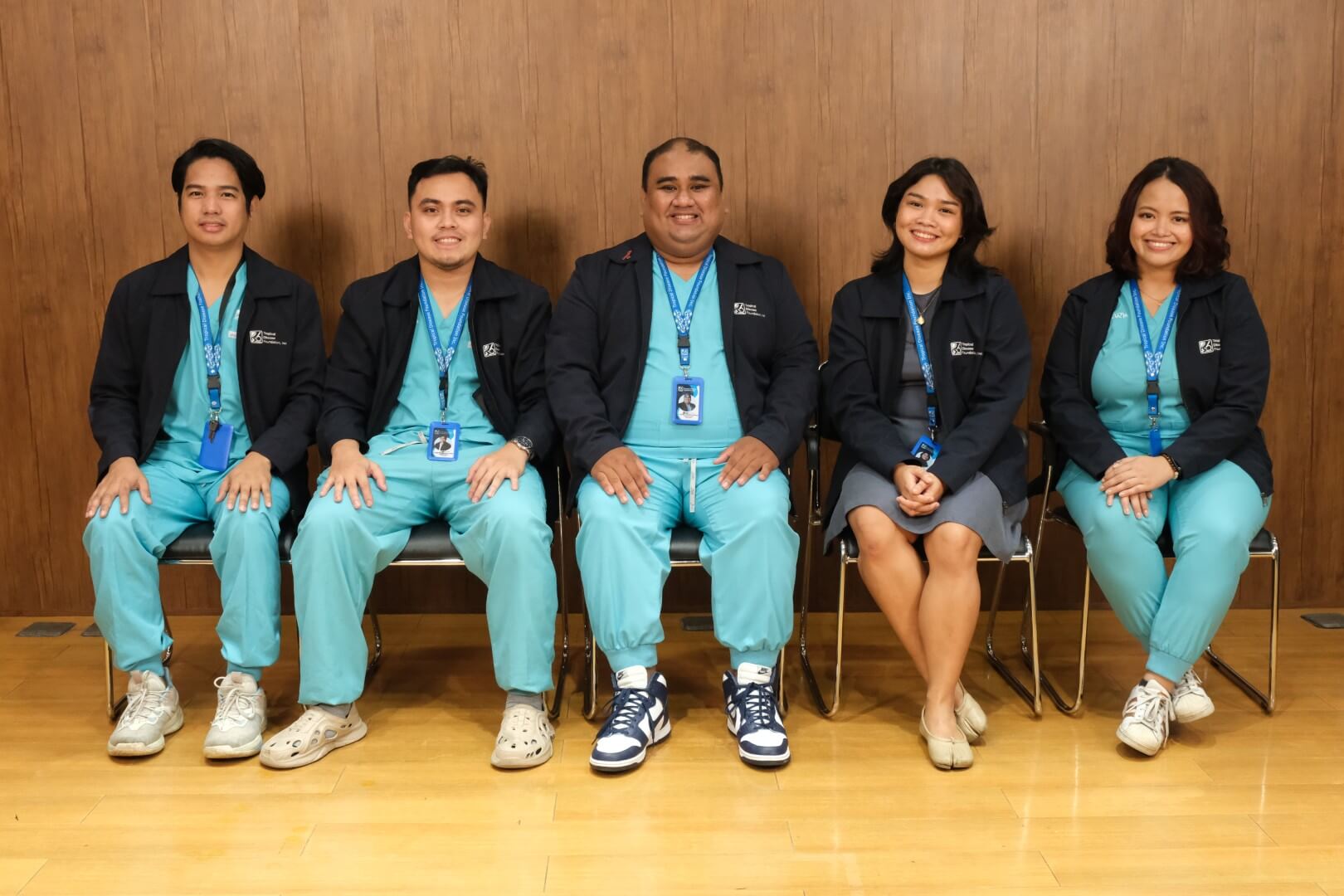 Four medical professionals sit in a row against a wooden backdrop. Three individuals wear light blue scrubs and dark jackets, while a woman on the right wears civilian attire.