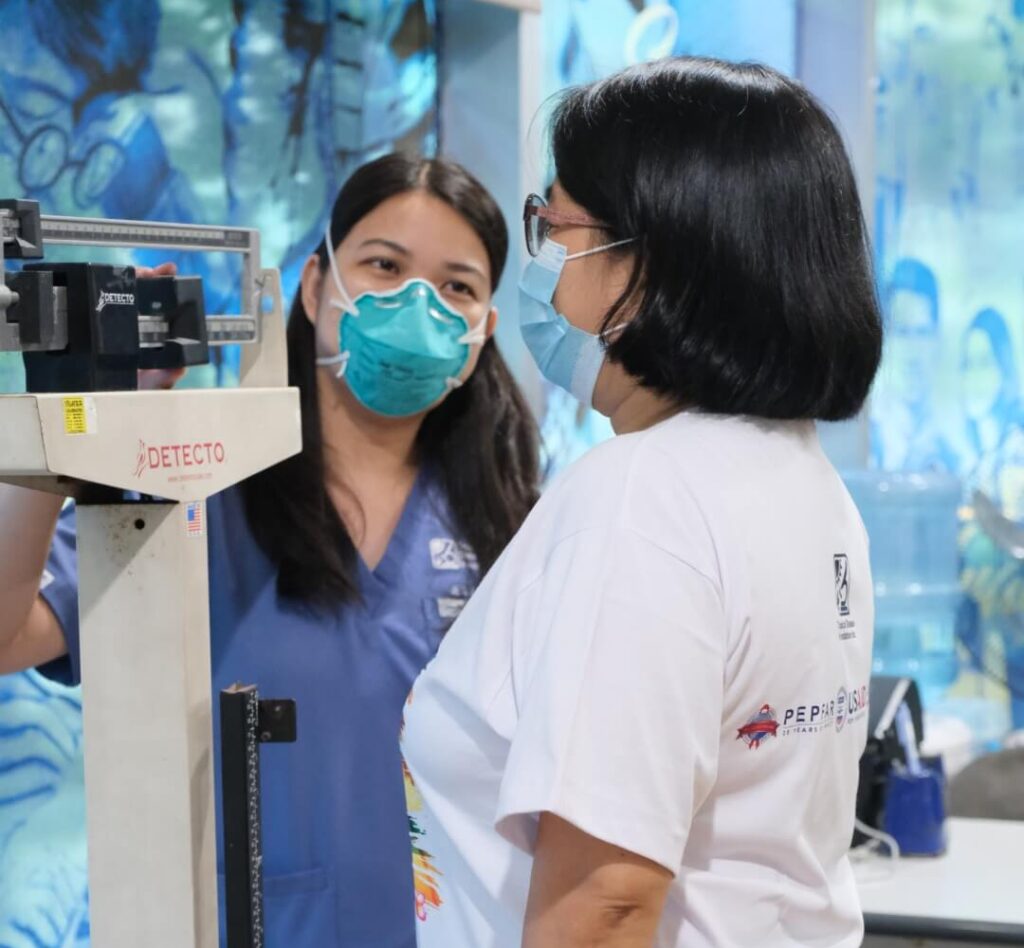 Two female healthcare professionals wearing protective teal face masks stand close together while reviewing medical charts and discussing critical patient treatment details inside a bright, modern hospital clinic environment.