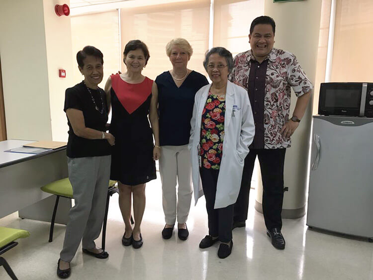 Five medical professionals and colleagues stand in a neat row inside a modern office corridor, smiling together for a group photograph while dressed in a mix of casual and clinical attire.