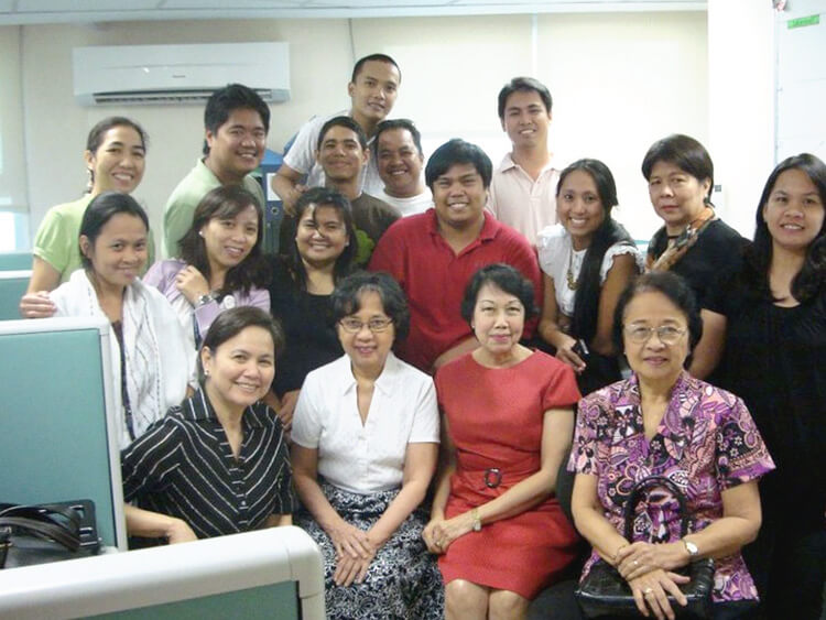 A cheerful indoor group photo shows a large gathering of healthcare staff and administrative colleagues posing closely together in multiple rows, smiling brightly for a commemorative office event picture.