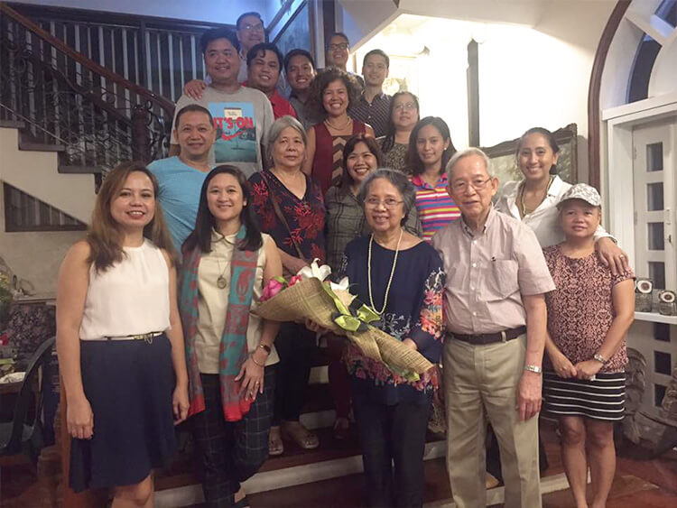 Gathered near an elegant staircase, a multigenerational group of family members and close colleagues smile warmly while presenting an elderly woman with a large, beautiful bouquet of flowers.