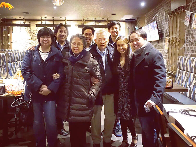 A joyous evening group photograph captures several friends and professional colleagues bundled up in winter jackets and coats, standing close together under warm, ambient indoor lighting.