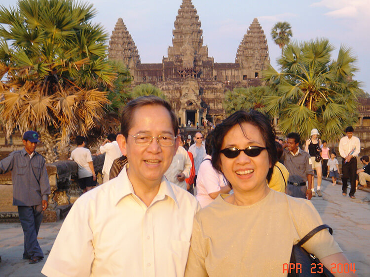 An outdoor travel photograph features a man and a woman smiling together in the foreground, with the historic stone towers of the Angkor Wat temple complex rising majestically behind them.