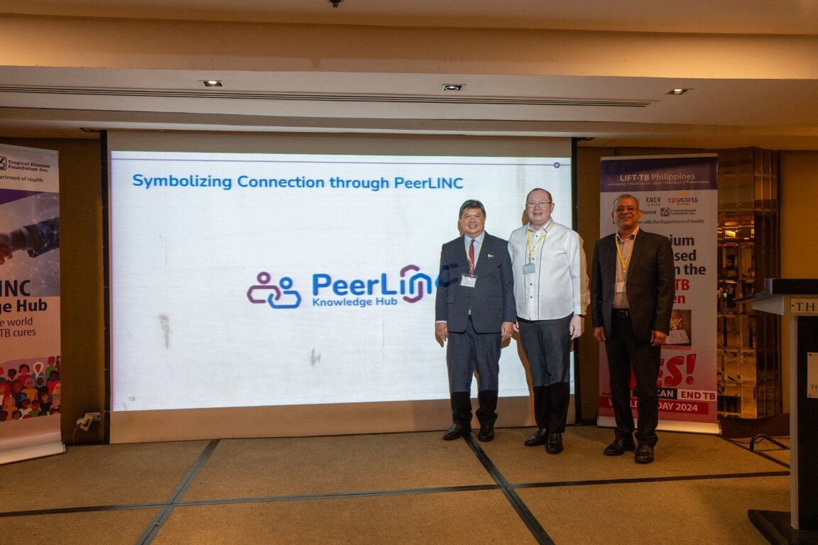 Dating from a 2024 event, this indoor photograph captures professional colleagues standing together on a stage next to a corporate backdrop displaying organizational branding during a formal conference presentation.
