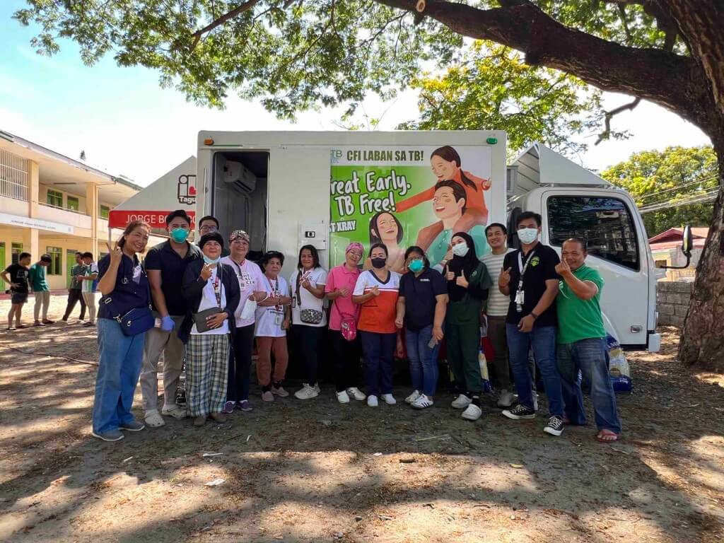 A large group of healthcare workers and volunteers pose together outdoors in front of a mobile health van during a community medical mission or public outreach event in 2025.