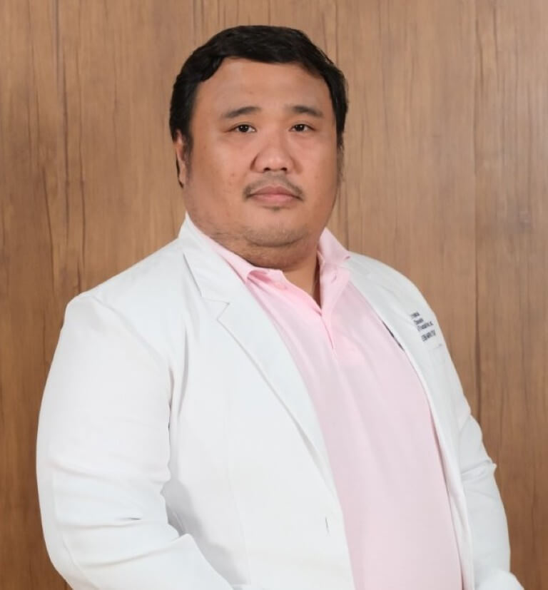 A formal medical headshot of Dr. Rholine Gem S. Veto looking forward thoughtfully, wearing a crisp white physician's coat over a light pink collared shirt against a wood backdrop.