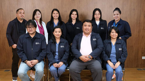 A group of ten medical professionals in matching blue jackets and scrubs pose together in a studio setting with a light brown paneled background.