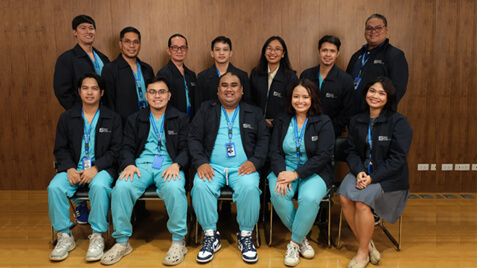 A group of twelve people, six men and six women, pose for a group photo, standing and seated in two rows against a wood-paneled wall.