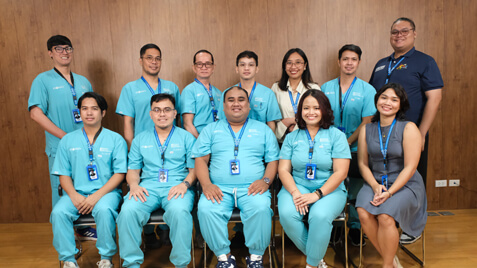 Group portrait of ten medical professionals in blue scrubs and two people in business casual attire smiling for the camera in front of a wood-paneled wall.
