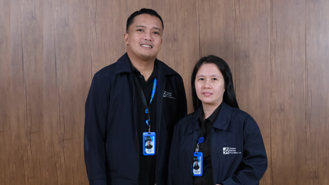 Two smiling employees in matching dark blue jackets and ID badges stand against a wooden panel wall.