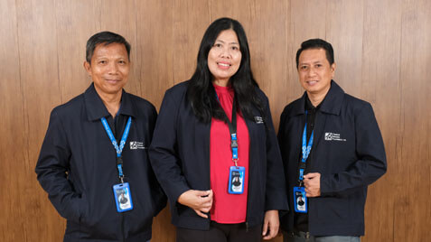 Three smiling people, two men and a woman, stand side-by-side against a wooden paneled wall, wearing matching navy blue jackets and employee lanyards with ID badges.