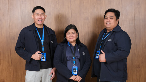 Three IT department employees in matching navy jackets and company ID lanyards pose against a textured wooden wall.