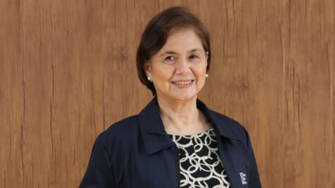 A smiling mature woman with short brown hair, wearing a patterned top and a dark jacket, stands against a textured wooden background.